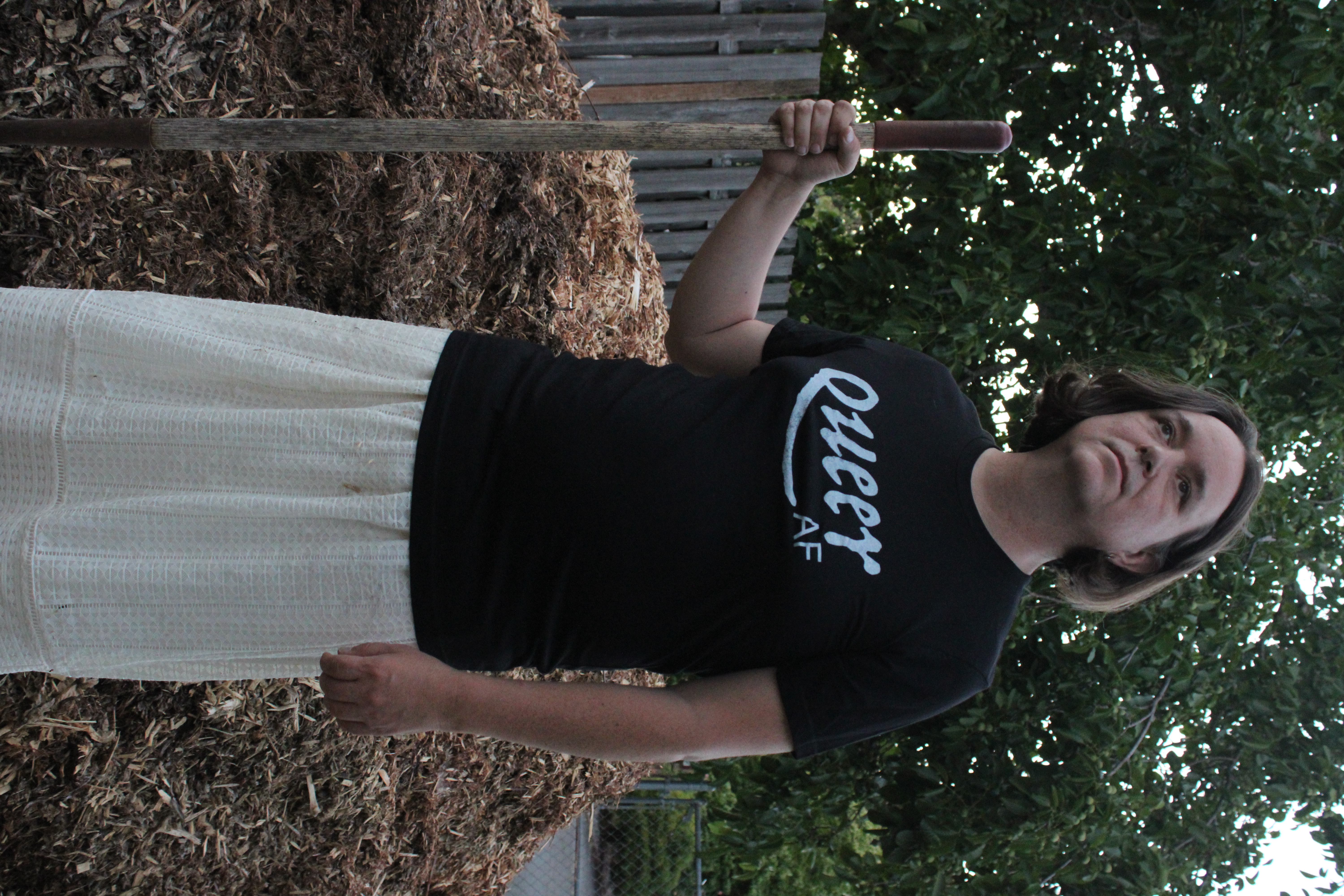 Jaelyn holding a shovel in front of a pile of wood chips wearing one of her shirt designs that reads "Queer AF"
